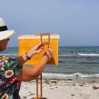 Frau mit Hut steht am Meer malt Pleinair und hält ein Passepartout